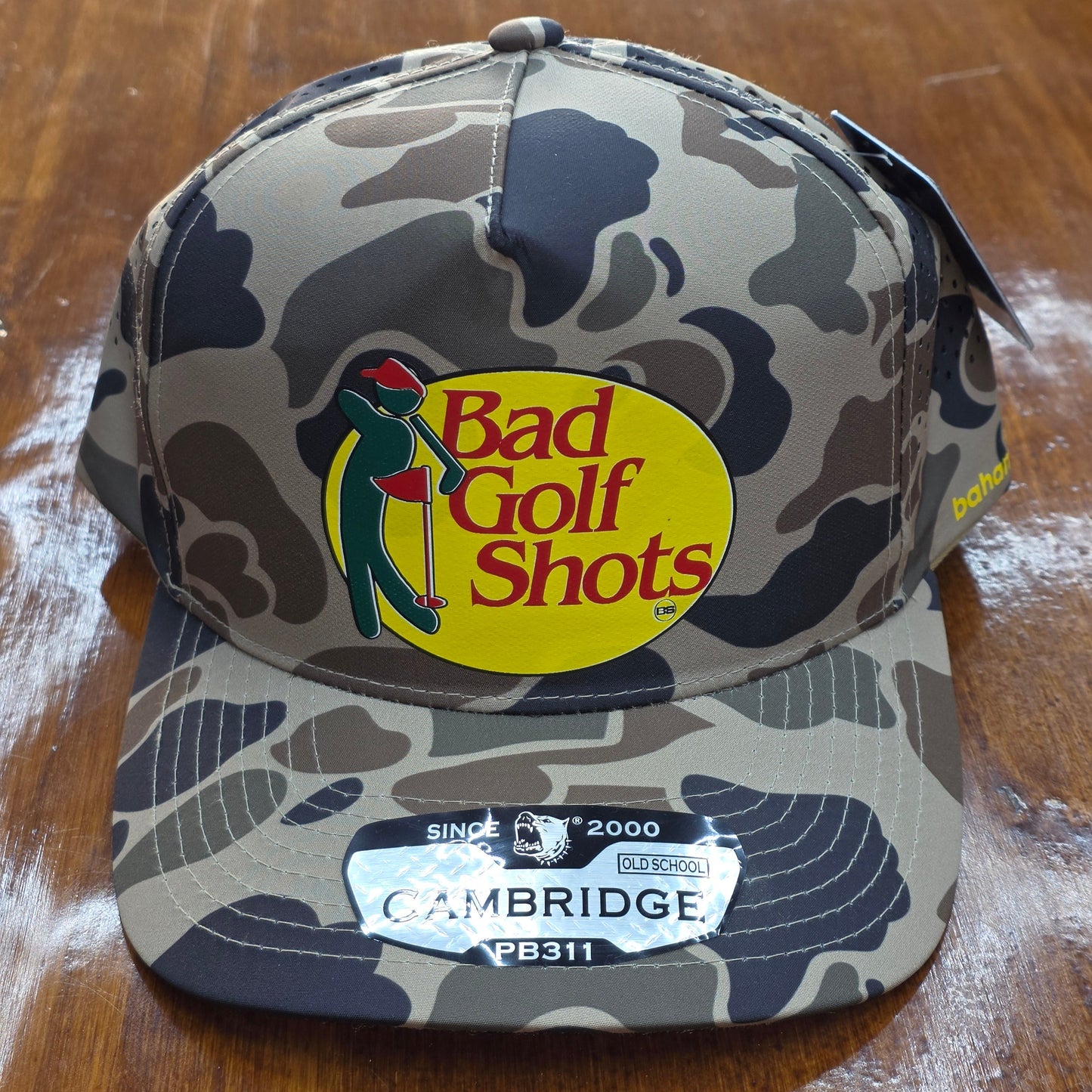 Camouflage golf ball case with 'Bad Golf Shots' logo on a wooden surface