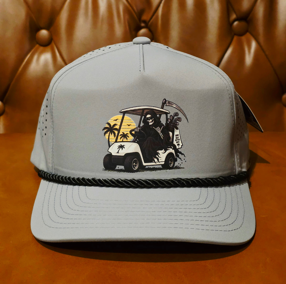 A light gray snapback cap with a black rope detail around the brim and a graphic print of the Grim Reaper driving a golf cart with palm trees and a sunset on the front panel, set against a brown tufted leather background.