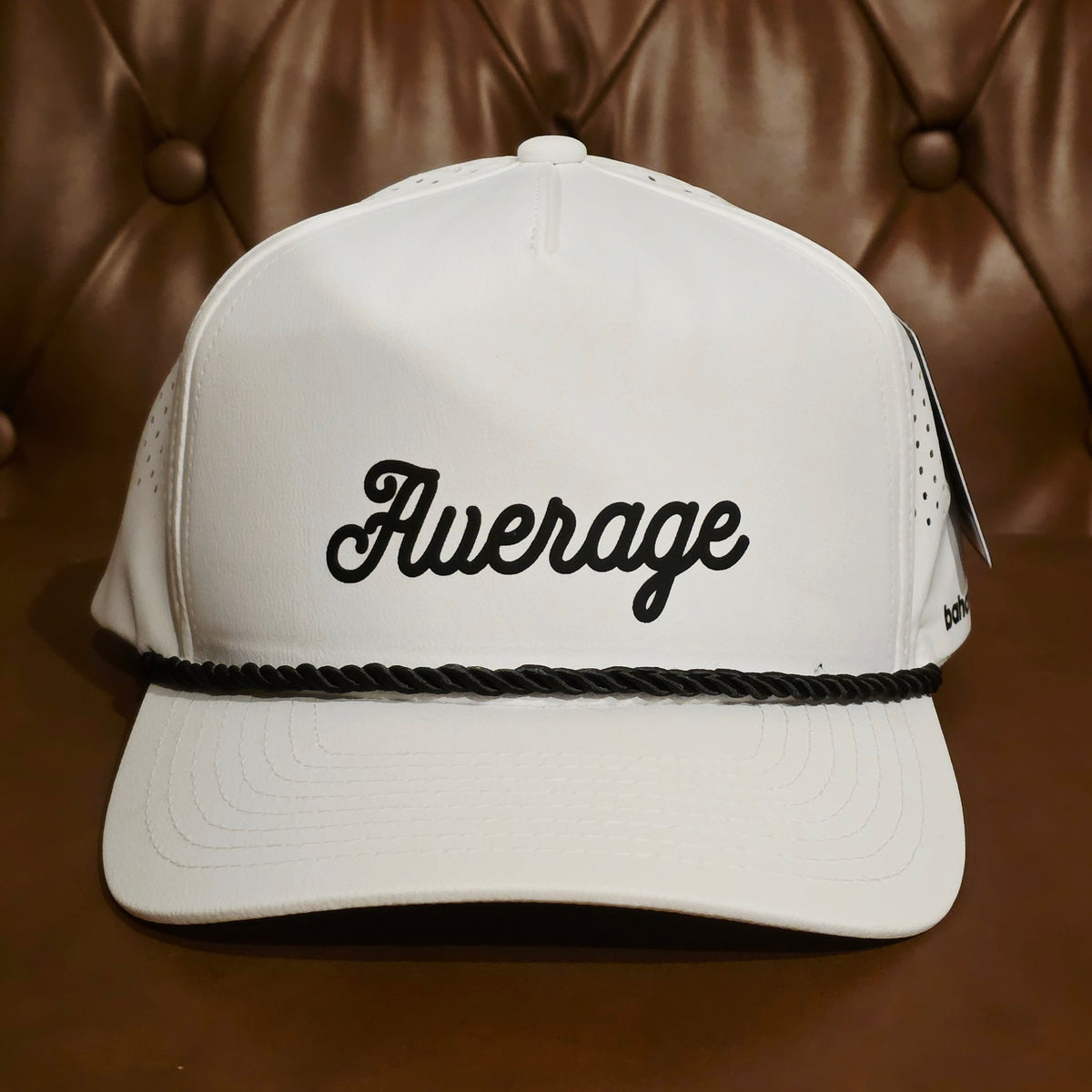 A white trucker-style baseball cap with black cursive embroidery reading 'Average' on the front, a black braided rope detail across the brim, and perforated side panels, photographed against a brown tufted leather background.