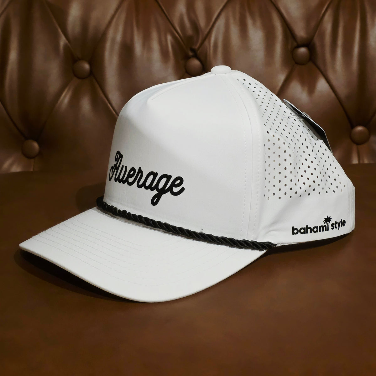 A white baseball cap with black embroidered script reading 'Average' on the front, black perforated side panels, a black braided trim across the brim, and 'bahami style' printed near the side. The cap is photographed on a brown leather tufted surface against a matching brown leather background.