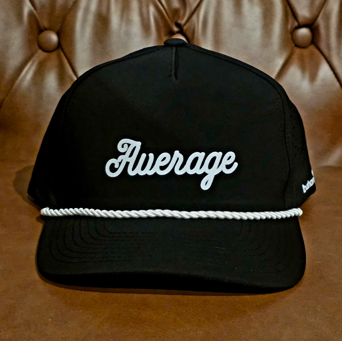 A black baseball cap with 'Average' embroidered in white script across the front, featuring a white braided rope detail across the brim, set against a brown tufted leather background.