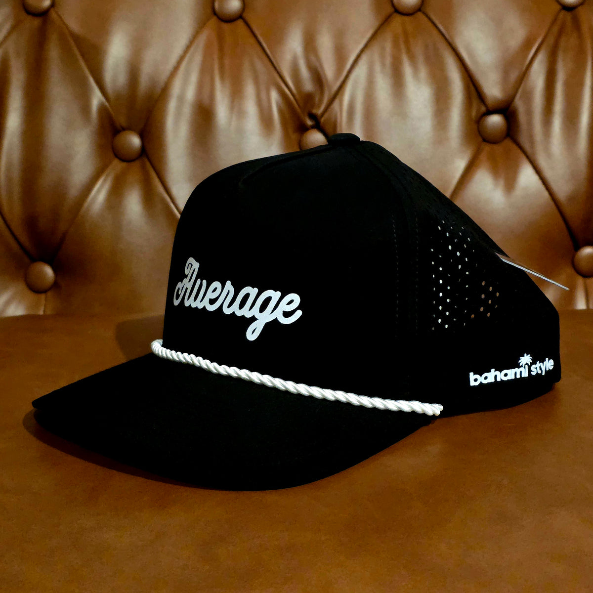 A black baseball cap with white script text reading 'Average' on the front and 'bahami style' on the side, featuring a white braided rope detail across the brim, set against a brown tufted leather background.