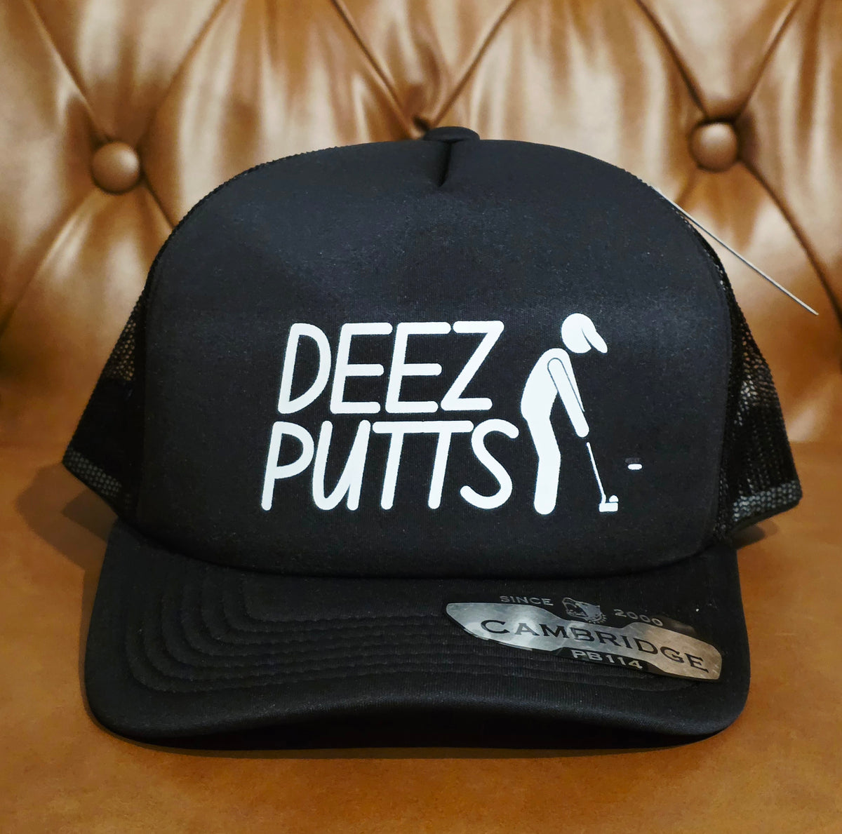 A black trucker hat with white text reading 'DEEZ PUTTS' and a white golfer graphic on the front panel, photographed against a tan tufted leather background. A Cambridge 1300 sticker is visible on the brim.
