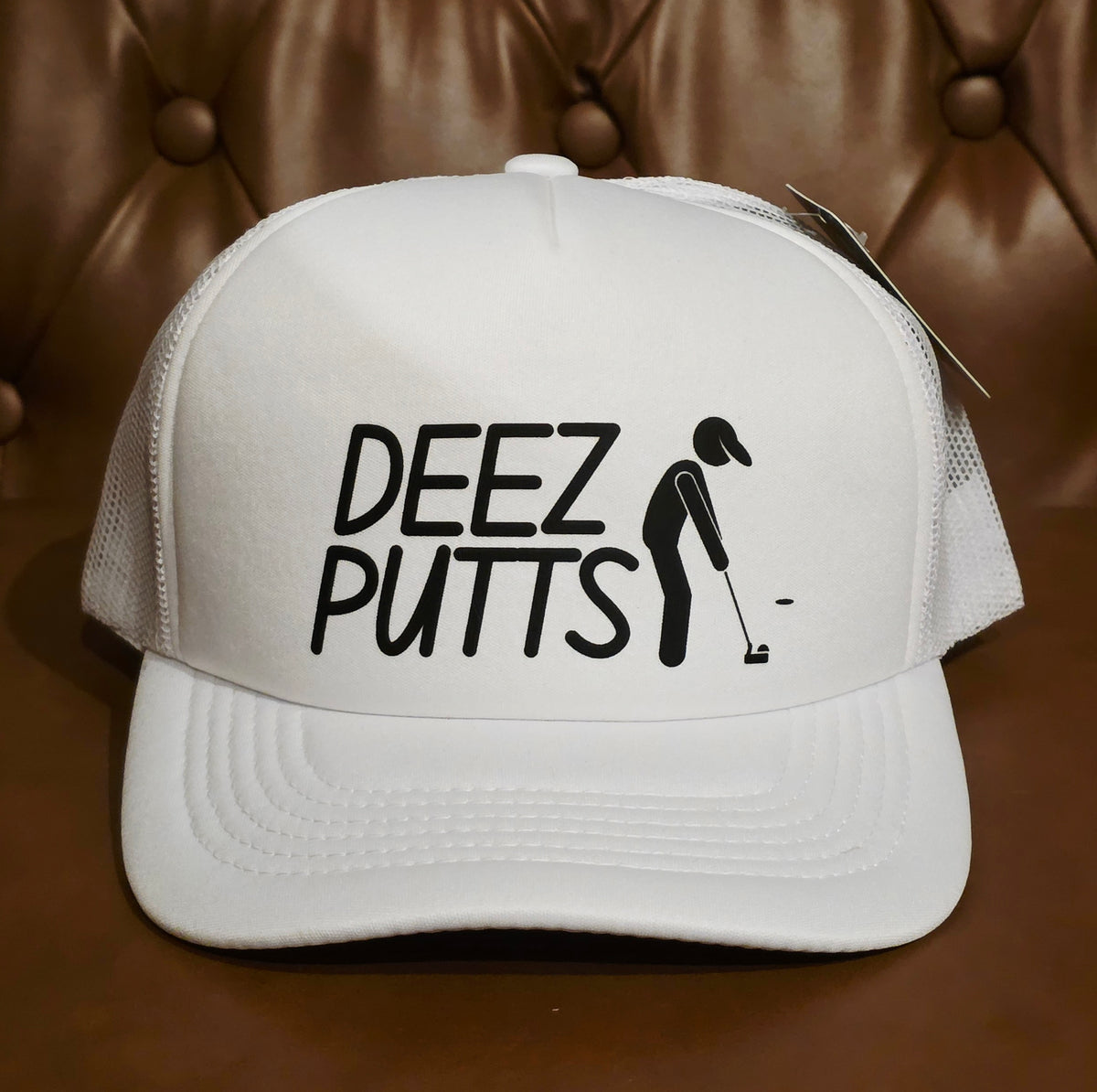 A white trucker-style baseball cap with black text reading 'DEEZ PUTTS' and a black golfer silhouette on the front, photographed against a brown tufted leather background.