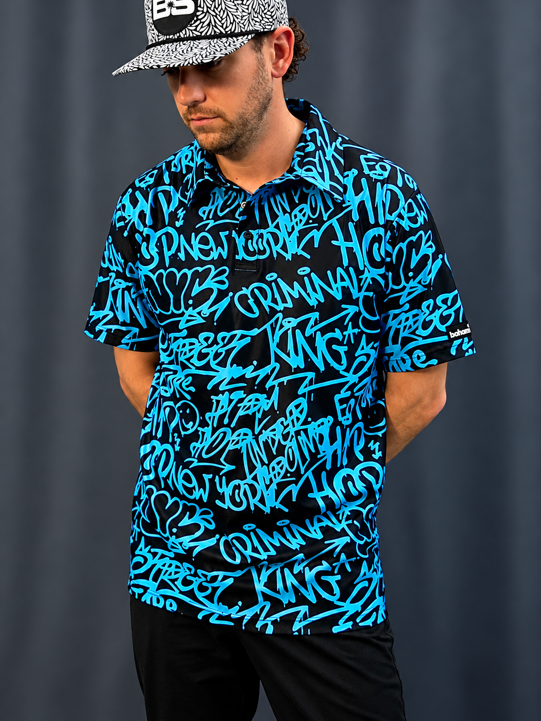 A man wearing a black short-sleeve collared shirt covered in bright blue graffiti-style script, paired with a black and white patterned baseball cap, standing against a dark gray curtain background.