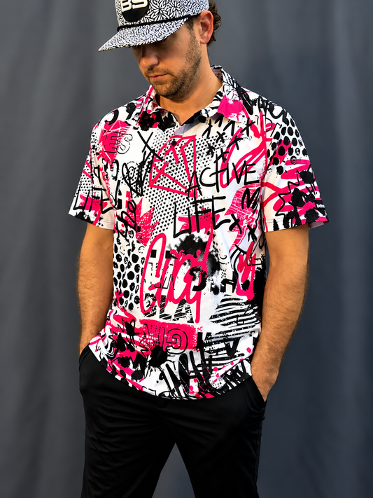 A man wearing a short-sleeve button-up shirt with a bold graffiti-style print in pink, black, and white, paired with black pants and a black-and-white patterned cap, standing against a dark gray backdrop.