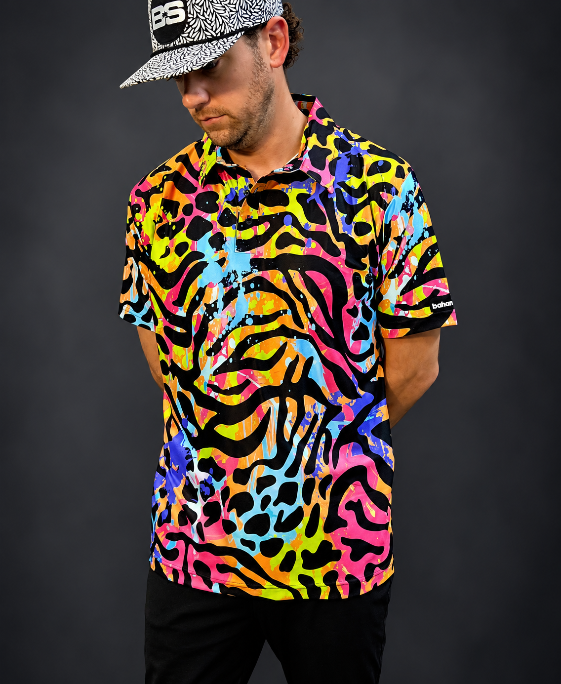 A man wearing a short-sleeve button-up shirt with a vibrant, multicolor abstract pattern in pink, yellow, blue, and black, paired with a black and white patterned baseball cap, set against a dark gray studio background.