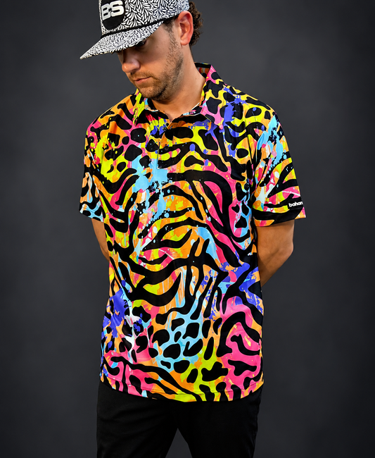 A man wearing a short-sleeve button-up shirt with a vibrant, multicolor abstract pattern in pink, yellow, blue, and black, paired with a black and white patterned baseball cap, set against a dark gray studio background.
